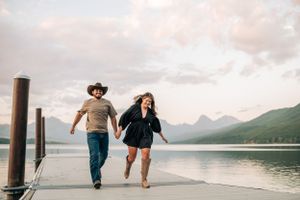Couple running joyfully hand-in-hand down wooden dock with mountain lake backdrop.