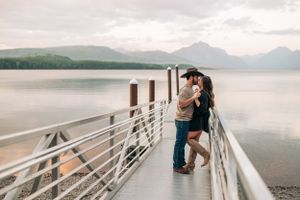 Intimate moment between couple on lakeside dock with mountains and cloudy sky.