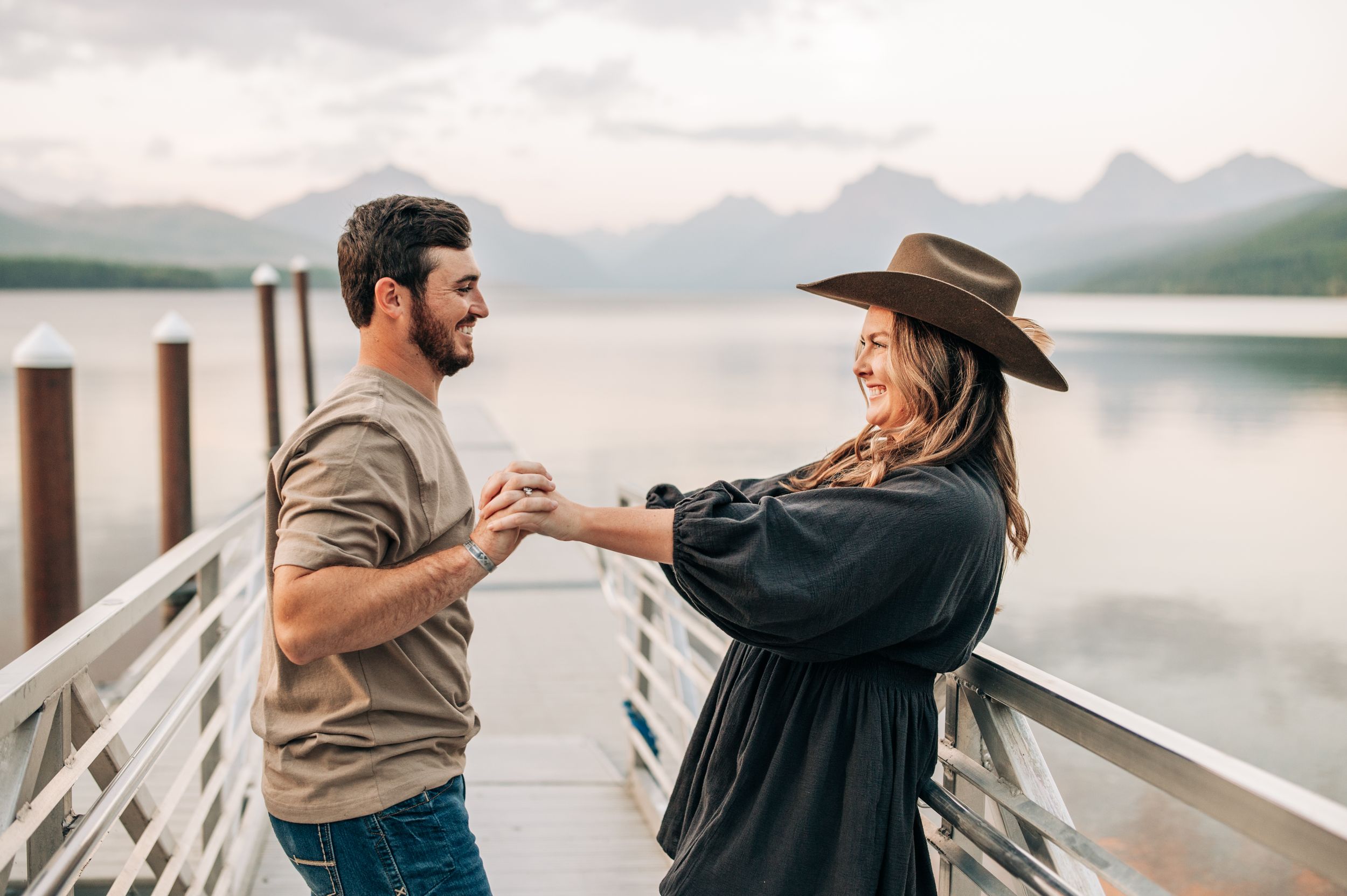 A couple dances playfully on a lakeside dock with mountains visible in the distance.