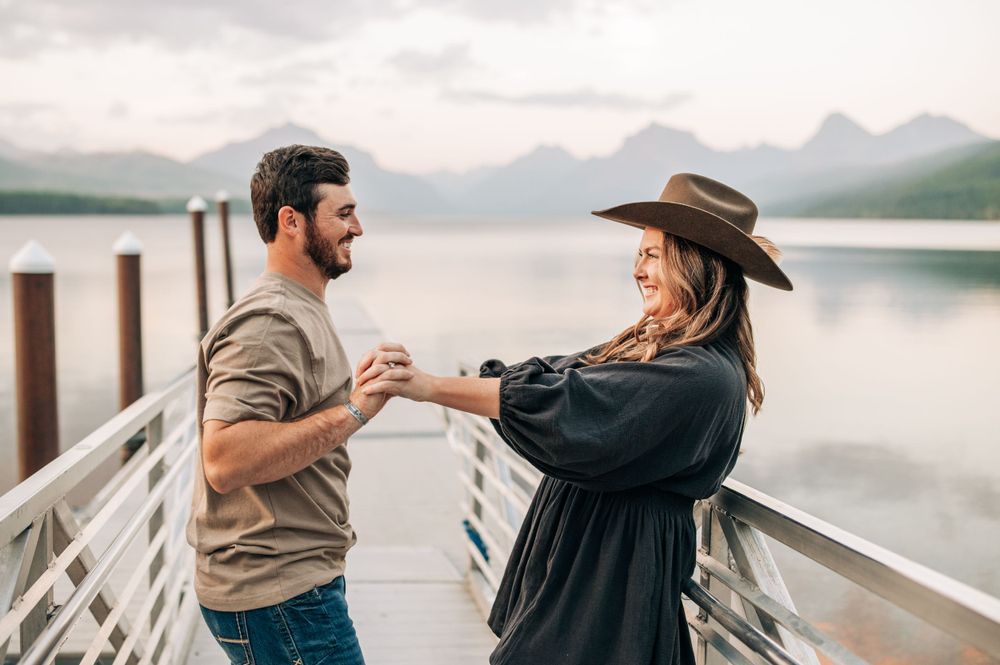 A couple dances playfully on a lakeside dock with mountains visible in the distance.