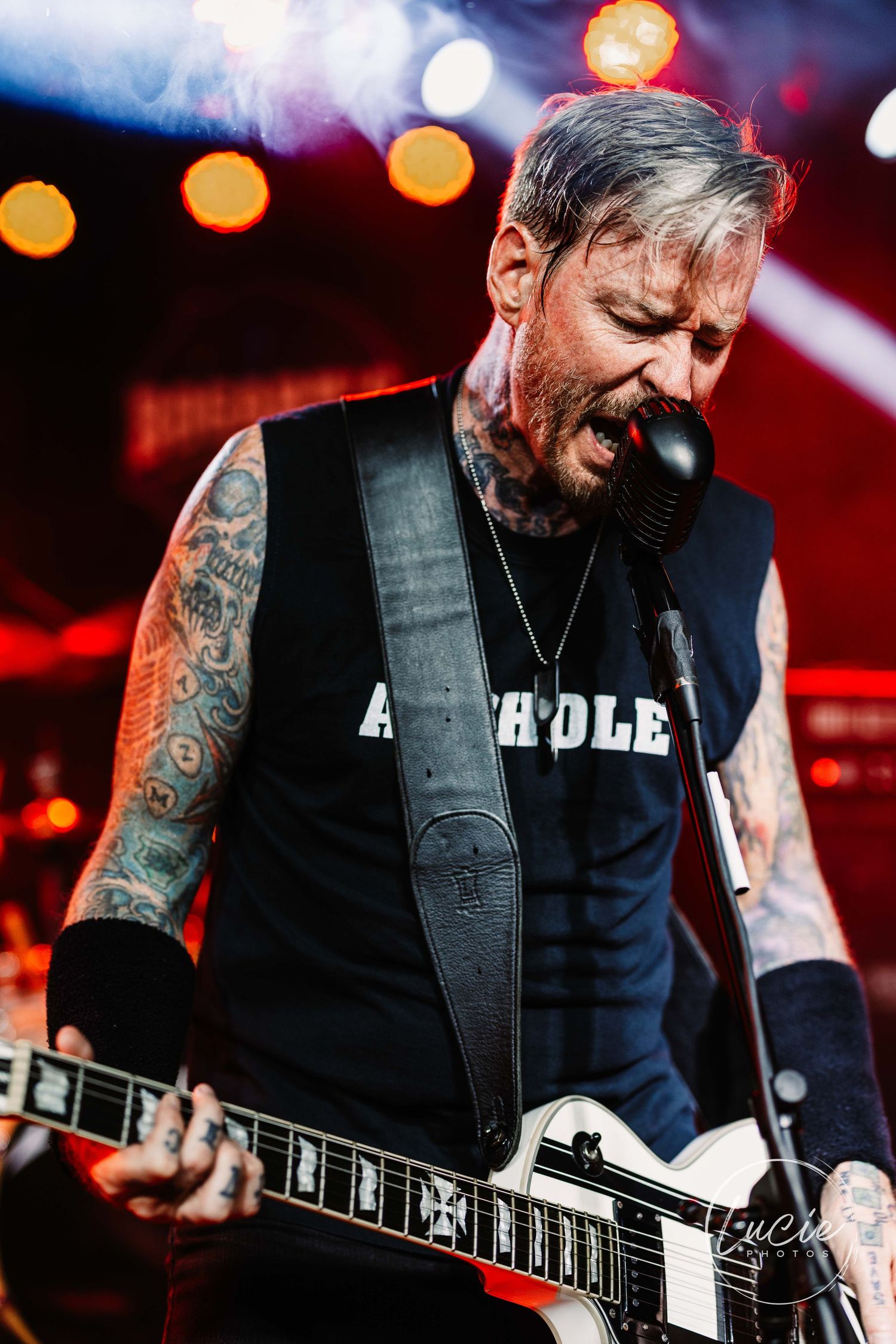 Guitarist with tattoos plays electric guitar on stage wearing black vest and sleeveless shirt under dramatic red lighting.