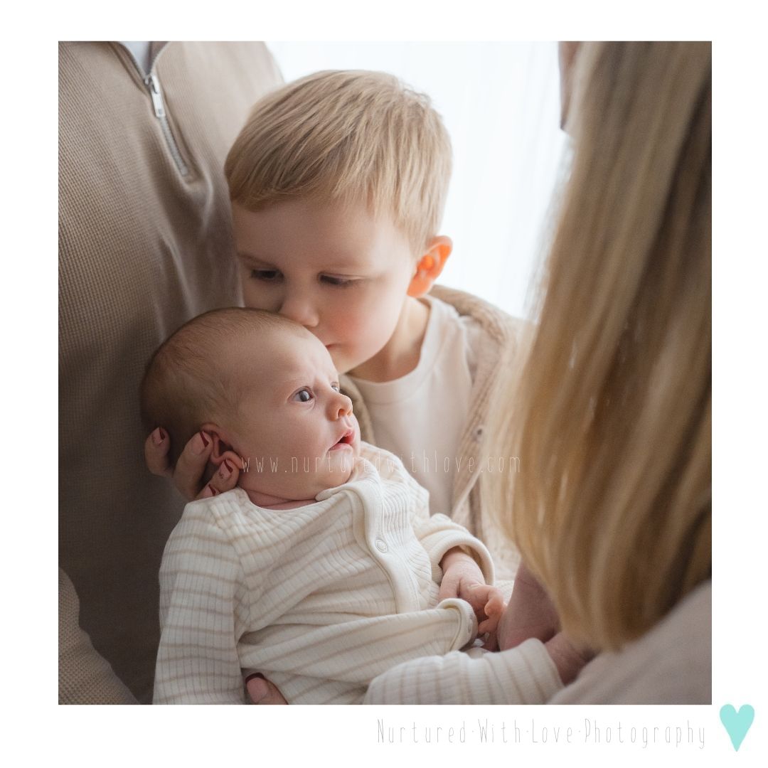 Toddler giving a gentle kiss on a baby's head while being held in soft natural lighting near window.