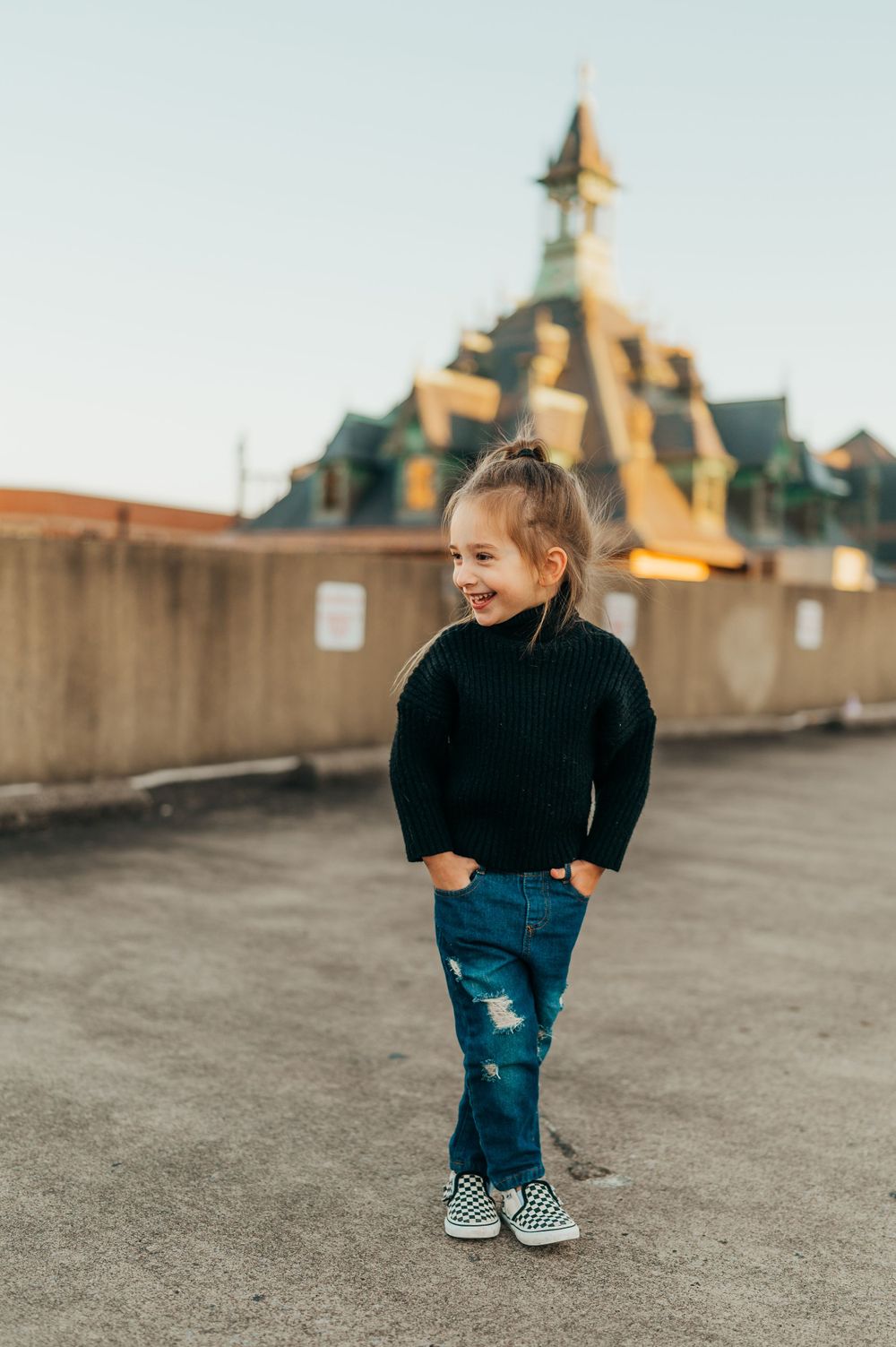 Small child wearing black sweater and ripped jeans poses on rooftop with architectural detail in background.