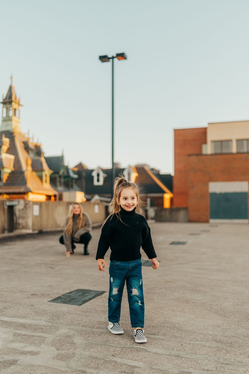 Child in black turtleneck and distressed jeans plays on rooftop with buildings in background.