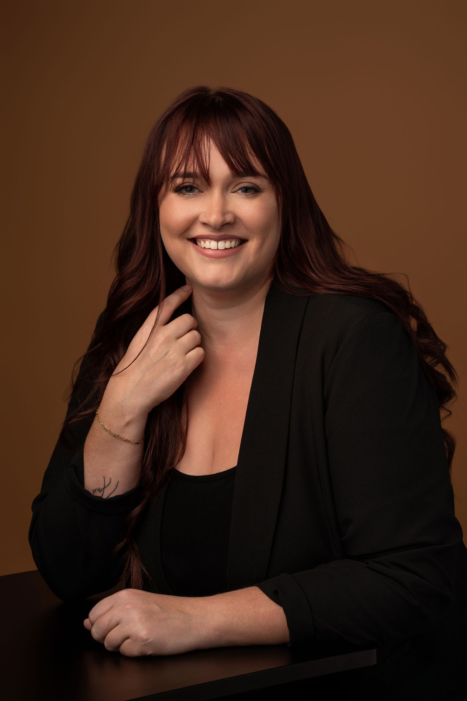 Smiling woman with red hair in black blazer poses confidently at a table against a warm brown background.