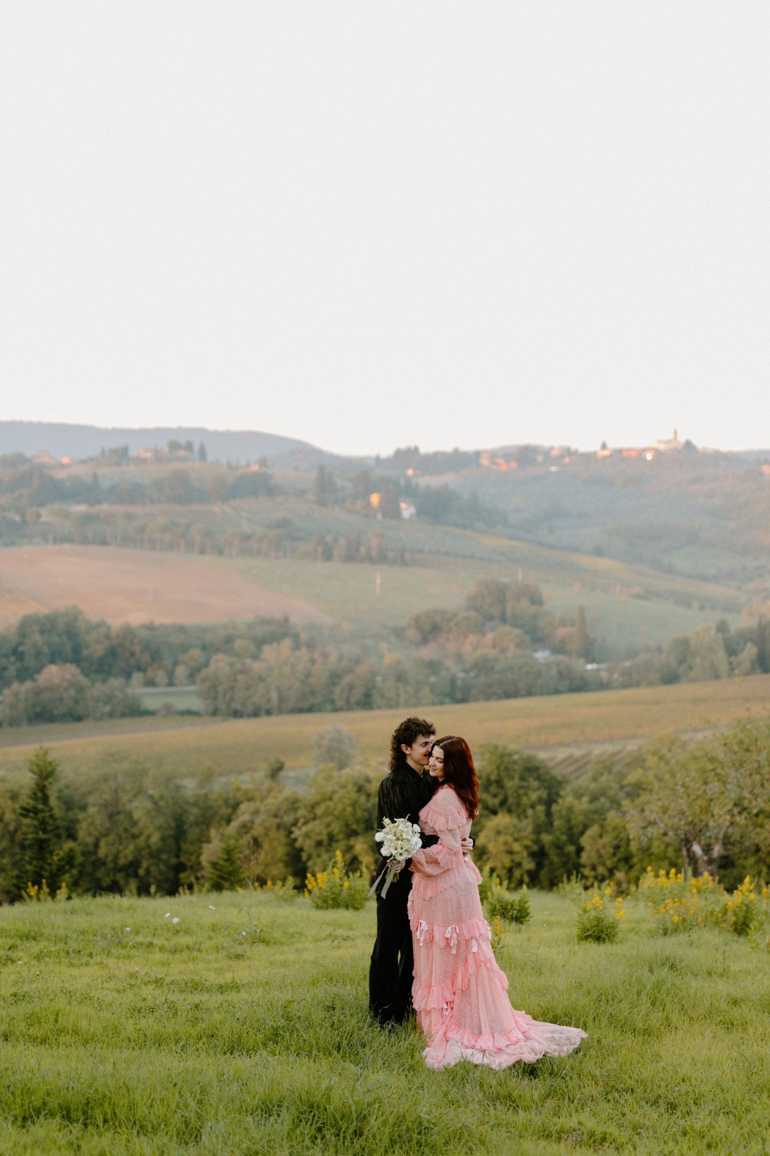 Couple embracing in pink ruffled gown and dark suit on lush green hillside with scenic Tuscan countryside backdrop.