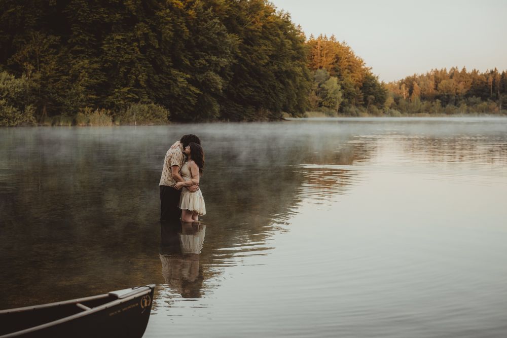 Two people embrace by a misty lake at sunset with a canoe in the foreground and autumn trees in the background.