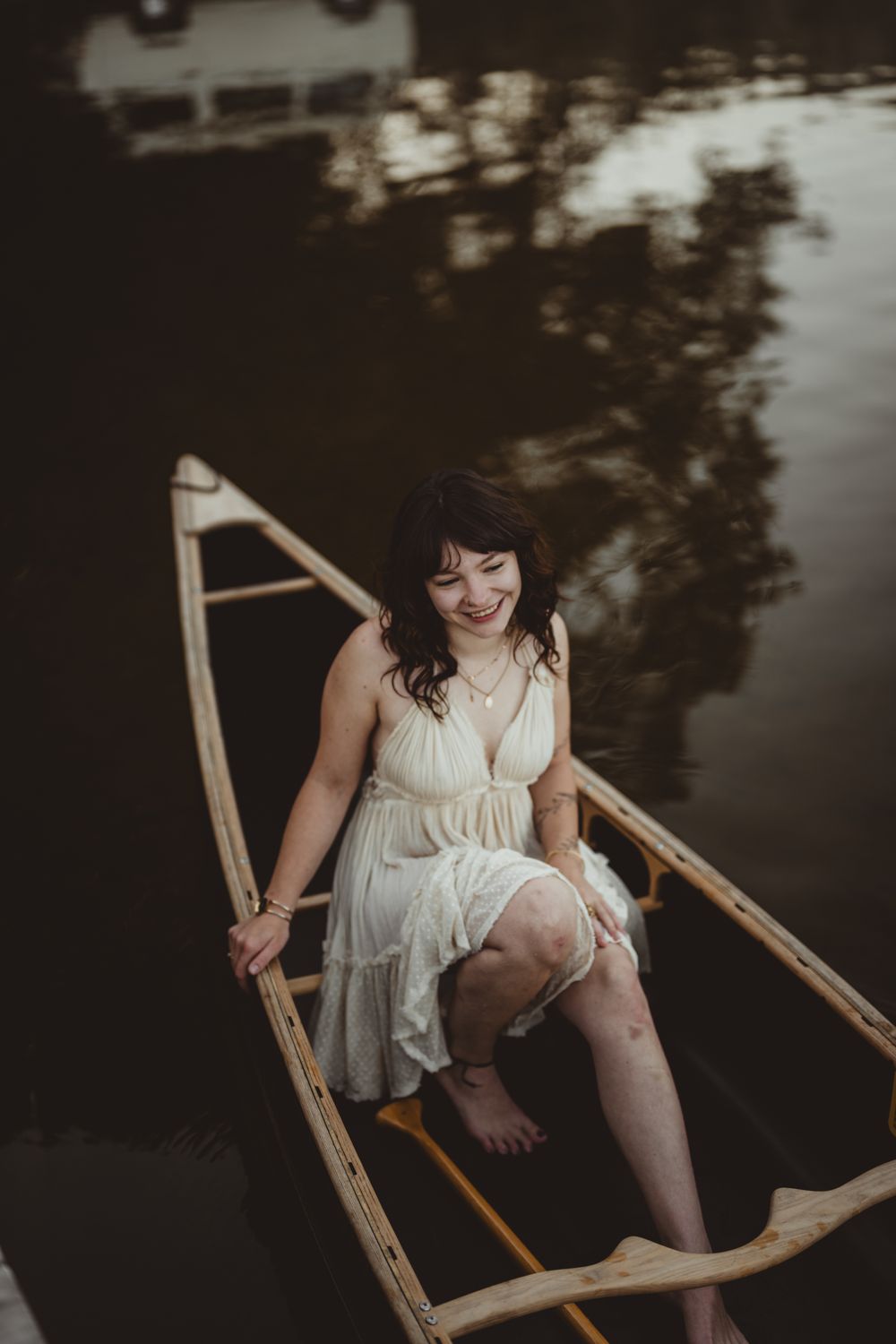 Person in white dress sits gracefully on wooden boat in dark moody water with reflective surroundings.