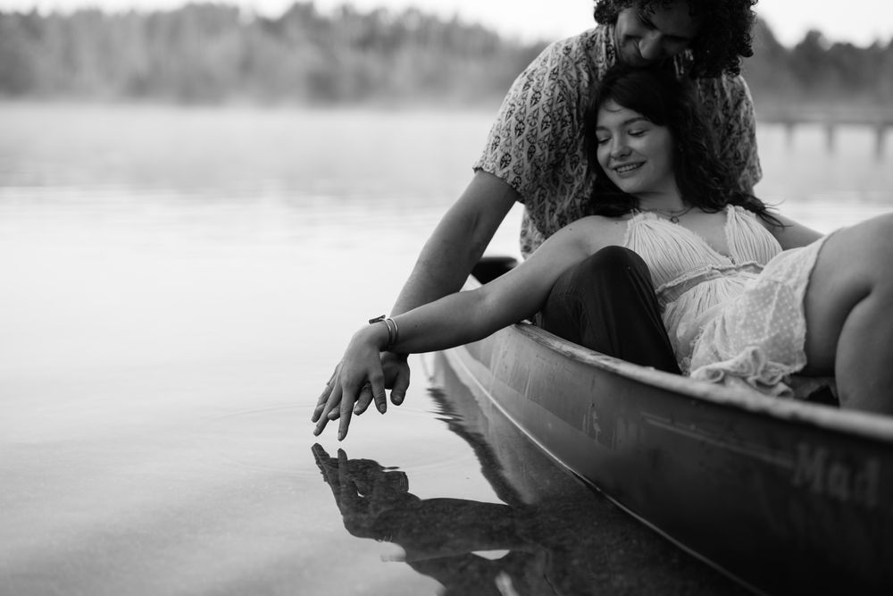 Black and white photo showing a couple lounging in a canoe on calm waters reaching to touch the lake's surface.