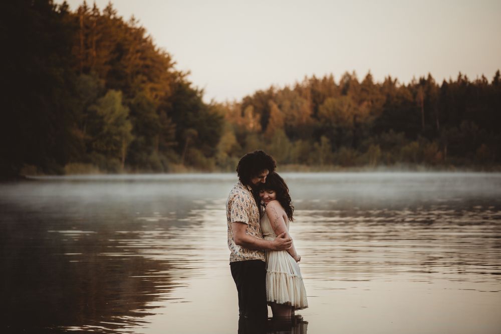 Two figures embrace in misty autumn lake with sunlit pine trees visible along the shoreline in golden evening light.