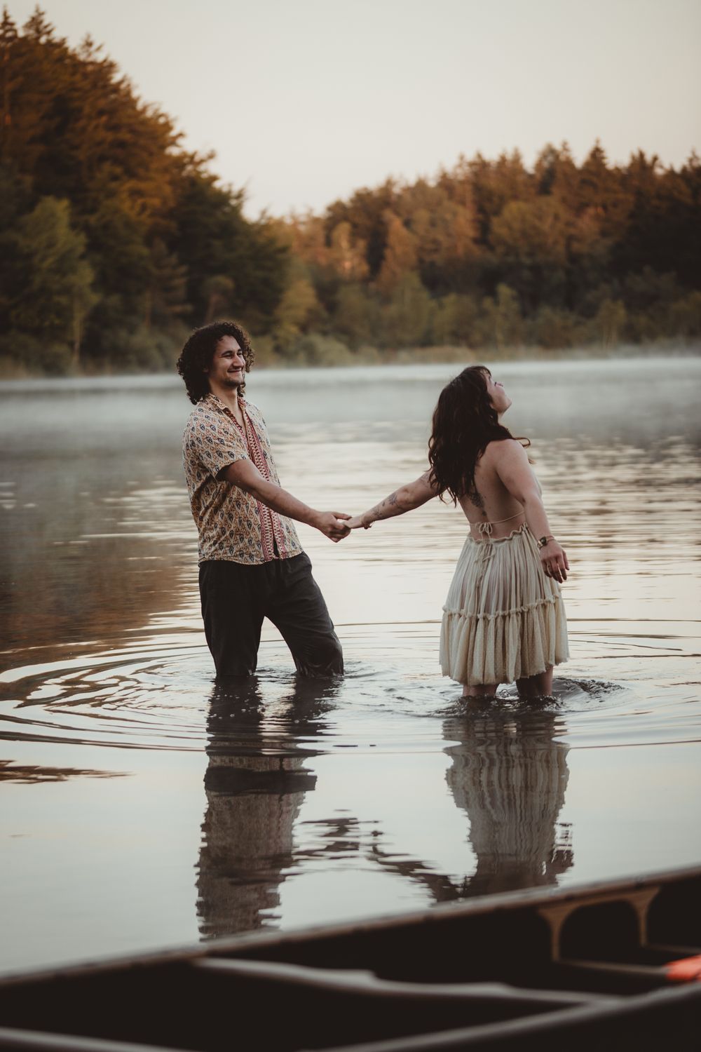 A couple stands together in shallow misty lake water at sunset, holding hands near an autumn forest.