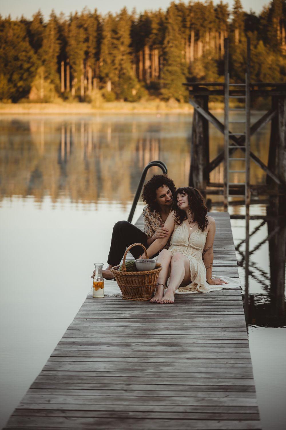 Romantic lakeside picnic on wooden dock with picnic basket and tranquil forest backdrop at sunset.