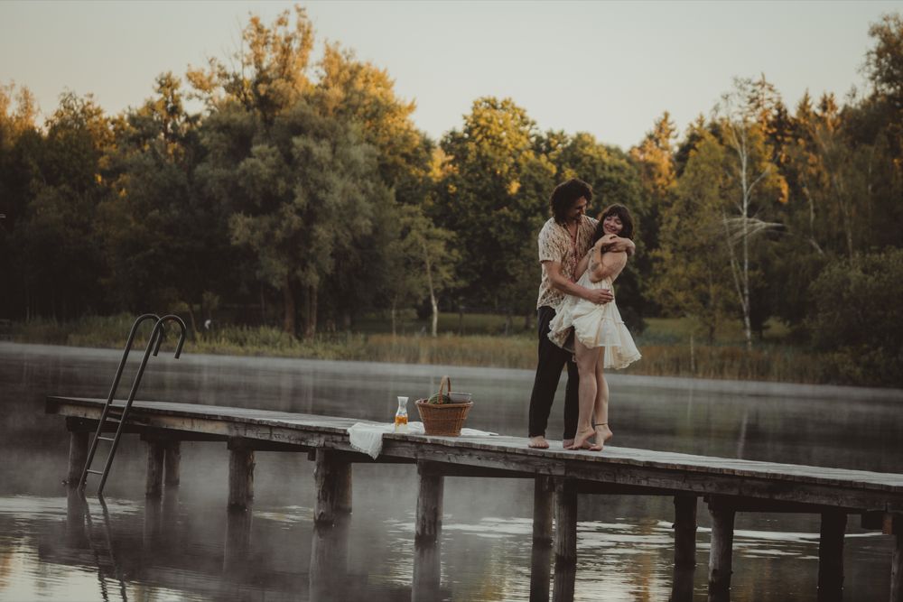 A romantic couple embraces on a wooden dock over a tranquil lake at sunset with golden light filtering through trees.