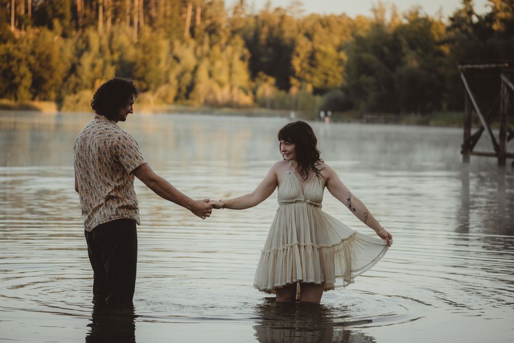 A romantic couple stands together in a lake at sunset, with her wearing a flowing cream dress and him in casual attire.