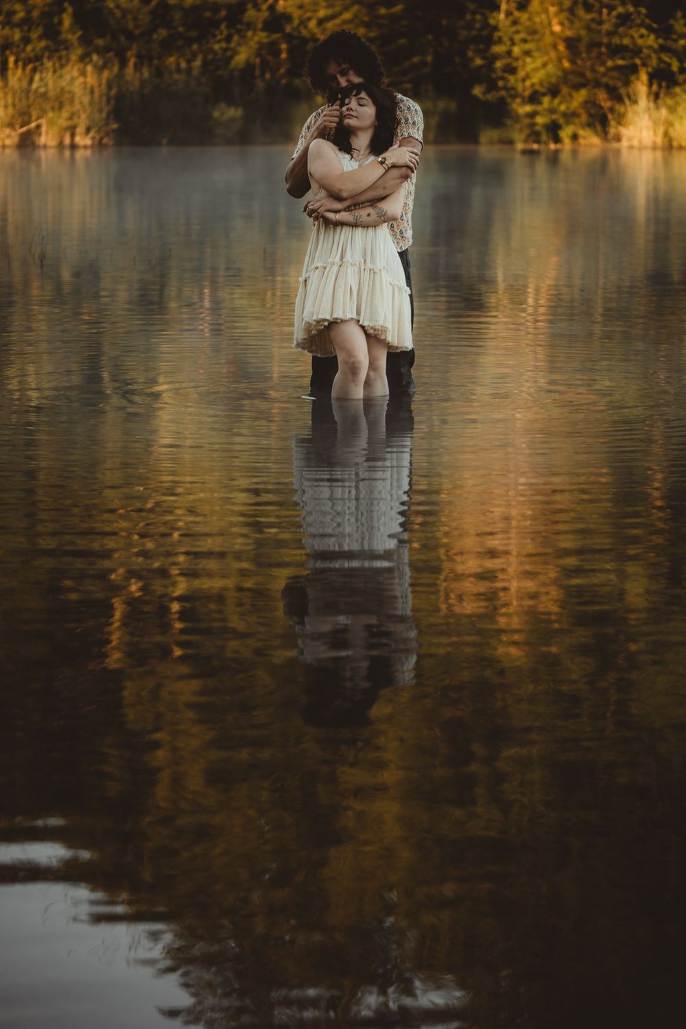 Dreamy romantic couple embraces while standing in reflective lake waters at golden hour.