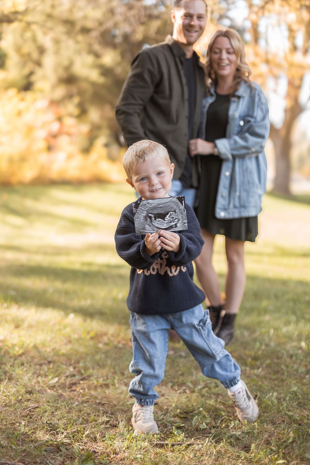 Toddler boy holding ultrasound photo in autumn park with smiling expecting parents in background.