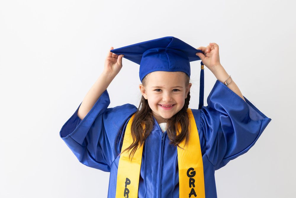 Smiling young girl in blue graduation cap and gown holding her mortarboard on white background.