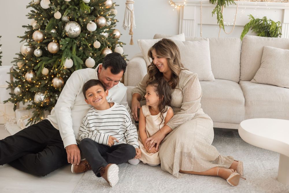 Happy family of four sitting together near a decorated Christmas tree in a bright living room.