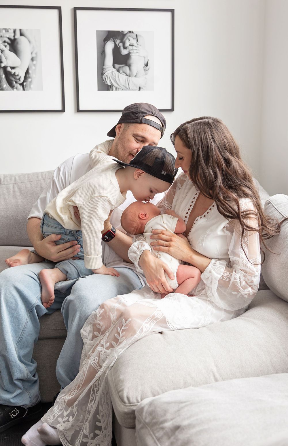 Parents and toddler lovingly gazing at newborn baby on a gray sofa in a modern living room.