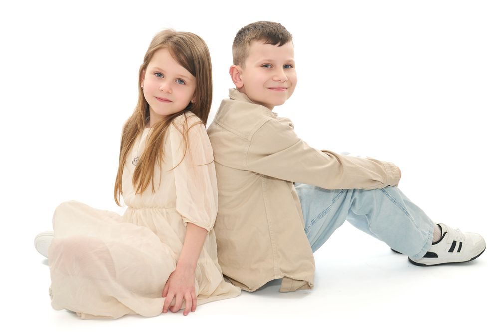 Two children sitting back-to-back on white background, girl in cream dress and boy in beige vest with jeans.