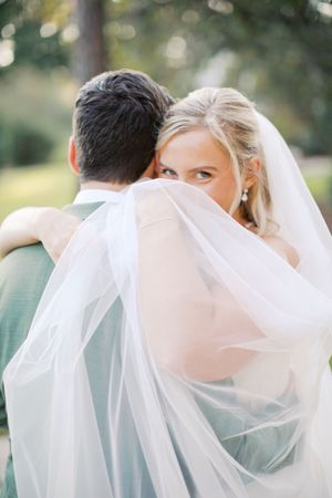 Bride peeks over groom's shoulder wrapped in flowing white veil during romantic outdoor wedding portrait at little river farms in milton ga