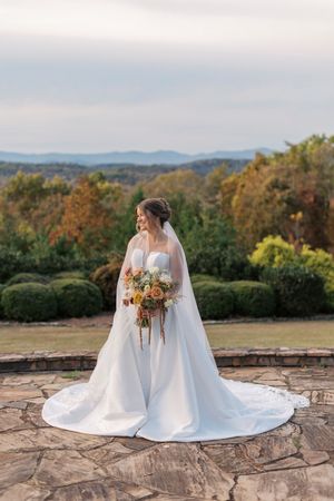 Bride in white ballgown and veil holds floral bouquet on stone terrace with mountain landscape backdrop at blue mountain vineyards