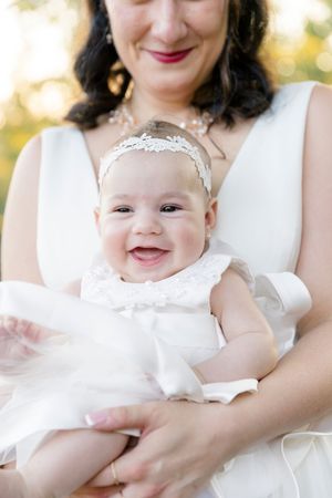 Smiling baby in white dress and headband held by mother in white dress outdoors at wolf mountain vineyards in dahlonega georgia