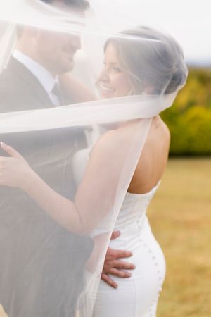 Bride and groom embracing outdoors, wrapped in a flowing white veil, smiling at each other on their wedding day at blue mountain vineyards
