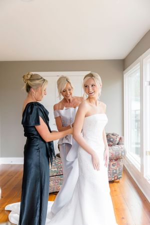 Bride smiling in white strapless gown assisted by two women in elegant dresses during wedding preparations at wolf mountain vineyards.