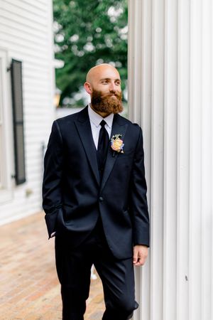 Bald groom with red beard in black tuxedo and floral boutonniere leans against white column at wedding at the carl house in auburn georgia