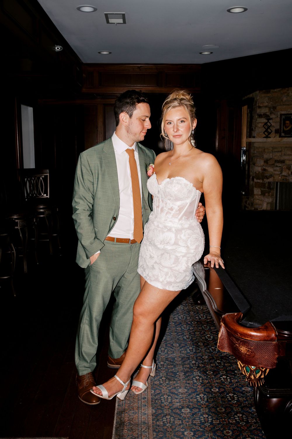 Bride in white mini dress and groom in sage green suit pose together at elegant indoor wedding at little river farms in milton ga