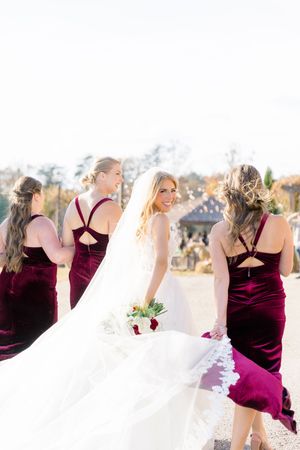 Bride in white gown holding bouquet smiles at camera surrounded by bridesmaids in burgundy velvet dresses at montaluce winery in dahlonega