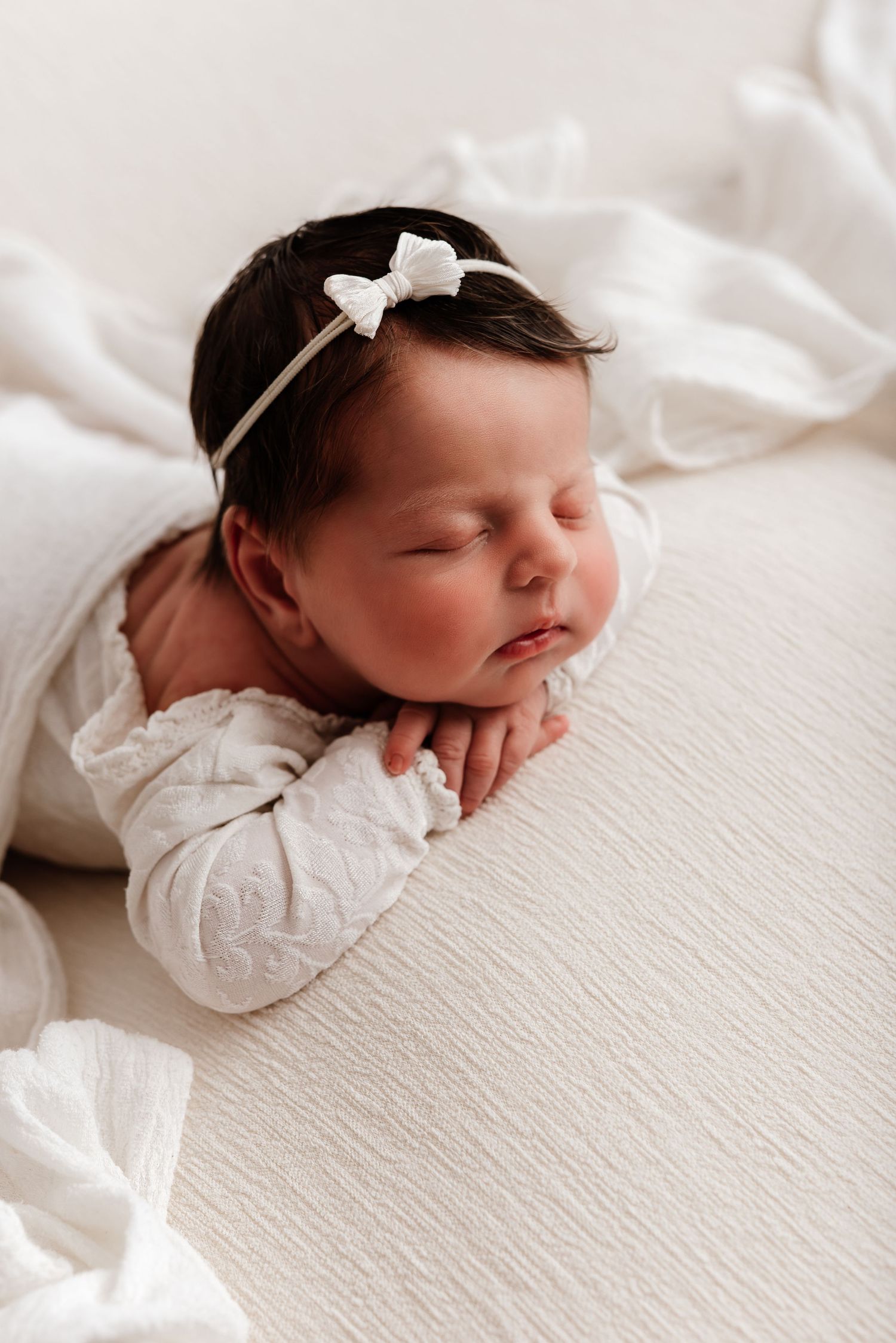 Sleeping newborn baby girl in white outfit and bow headband, peacefully resting on white blanket.