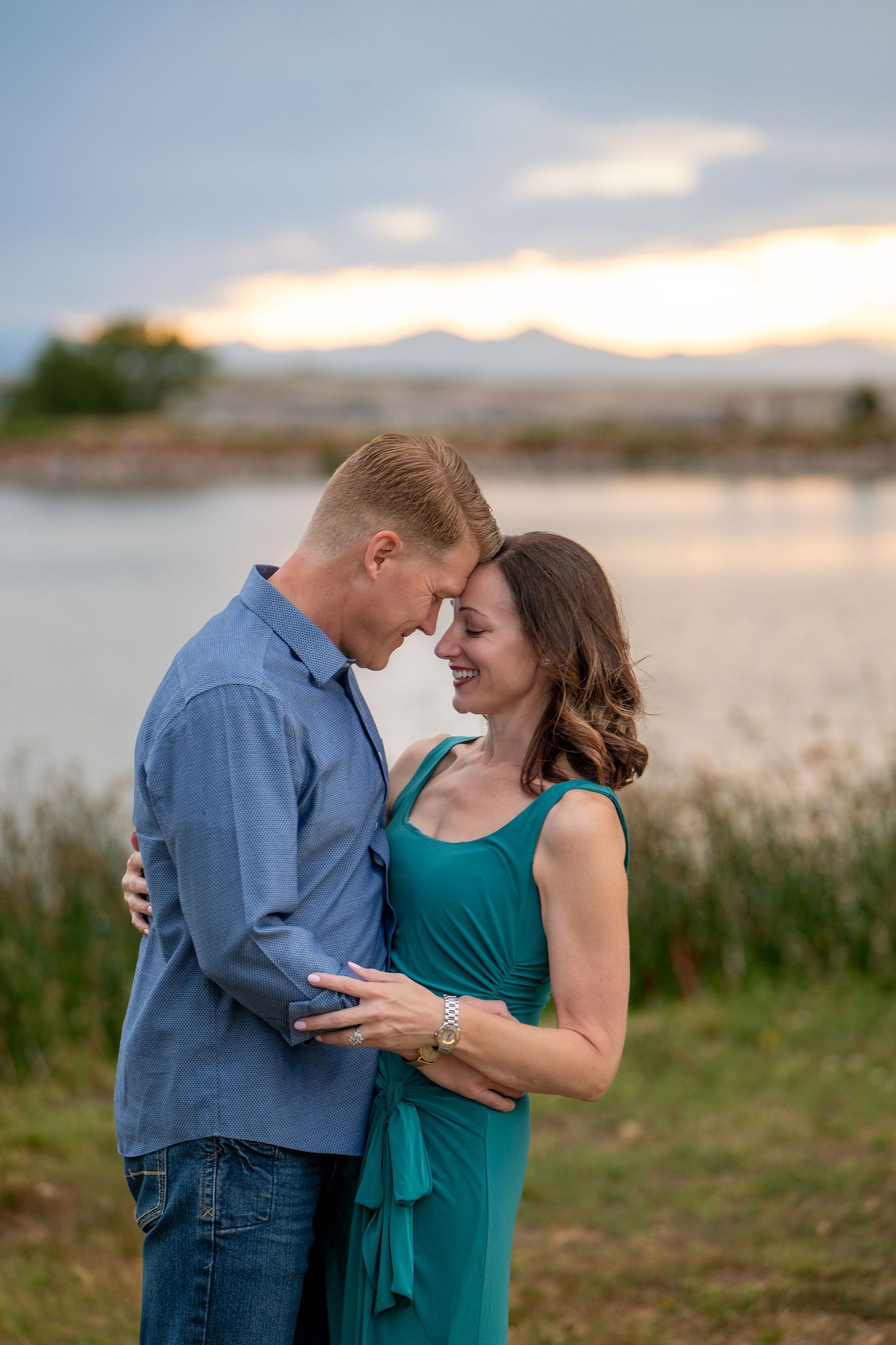 Couple in romantic pose by lake at sunset with mountains in background.