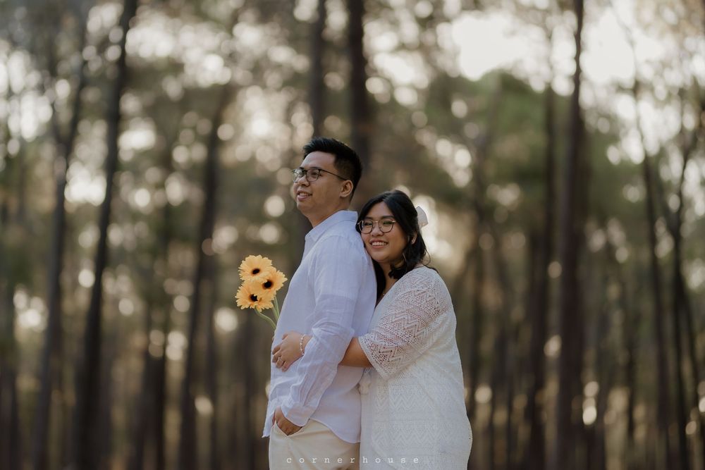 A couple in light colored outfits stand back to back in a dreamy forest setting during golden hour.