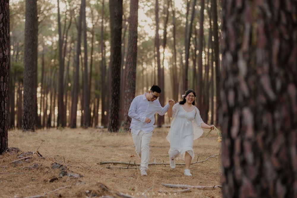 Two people in white outfits walk together through a sunlit forest with tall pine trees.