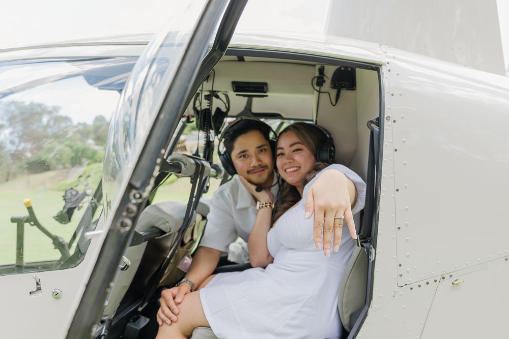 Smiling couple posing together in a helicopter cockpit while showing off an engagement ring.