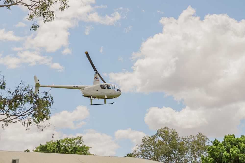 A white Robinson R44 helicopter hovers low over trees against a cloudy blue sky on a sunny day.