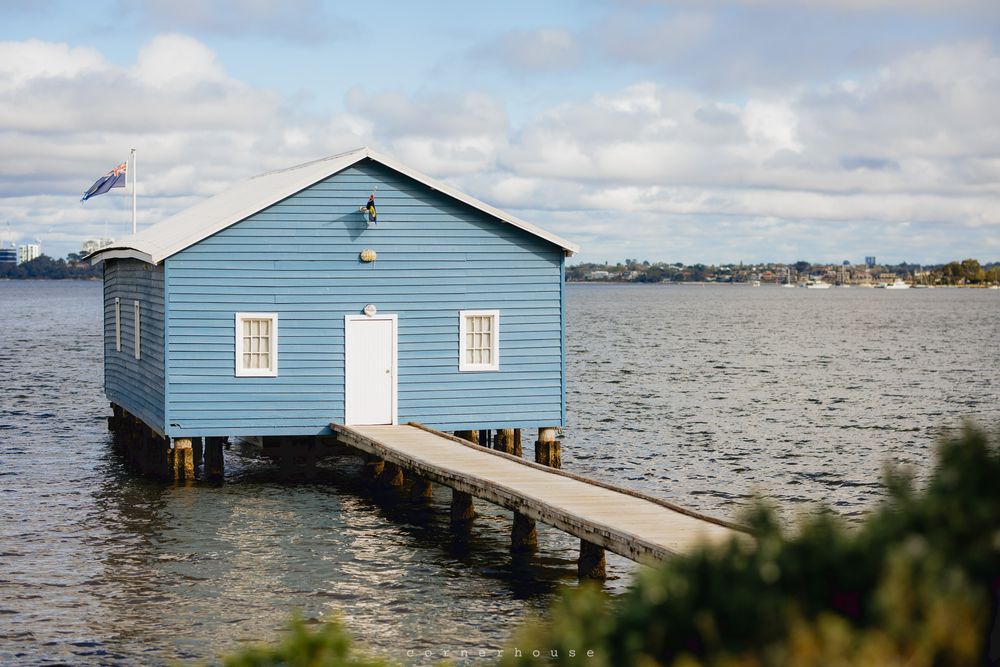 Iconic blue boathouse with white trim stands on wooden stilts over calm waters of Perth's Swan River.