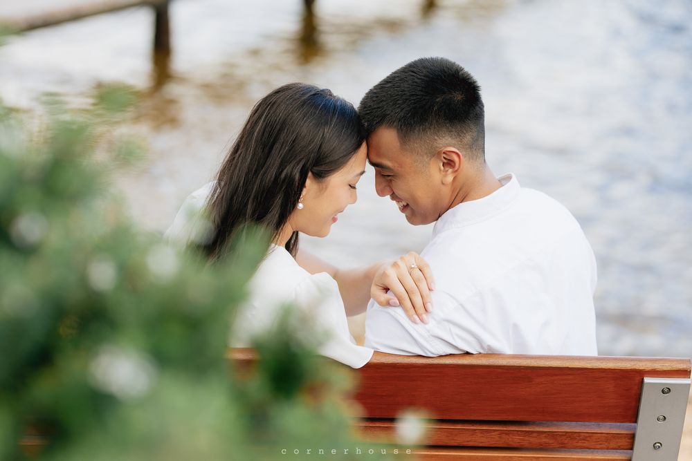 A romantic couple in white outfits share intimate moments while sitting on a wooden bench outdoors by water.