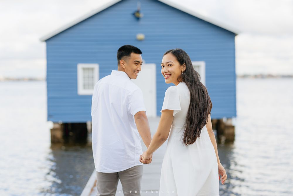 A couple in white outfits holding hands while standing together near a blue boathouse on the water.