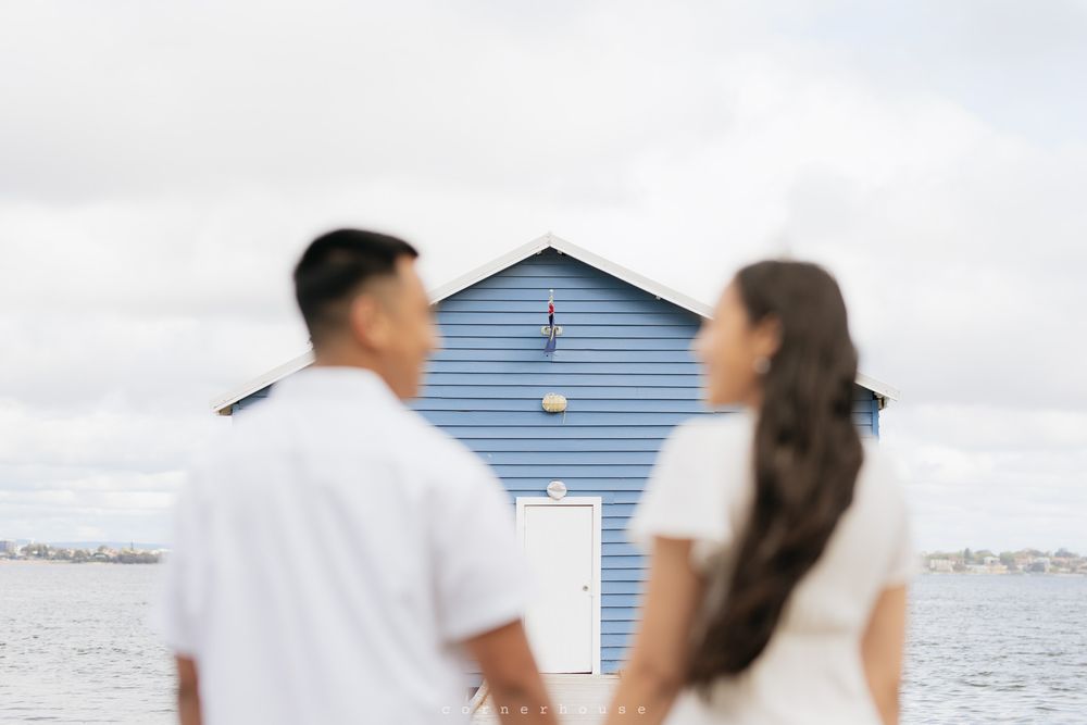 A couple stands blurred in the foreground with a blue and white beach hut against a seaside backdrop.