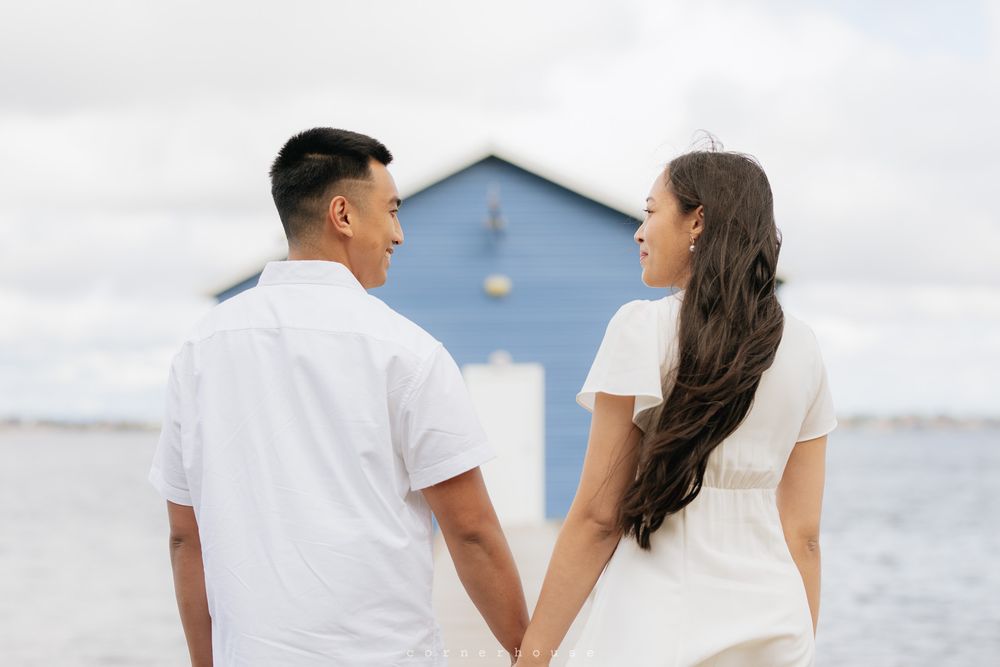 A couple in white outfits holds hands while standing by the ocean with a blue beach house in the background.