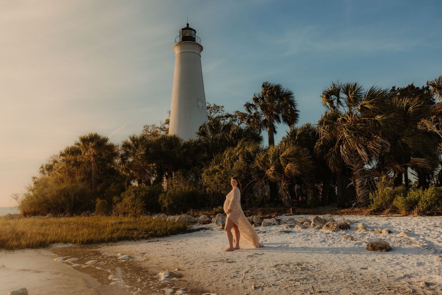 A towering white lighthouse stands amid palm trees on a sandy beach during golden hour.