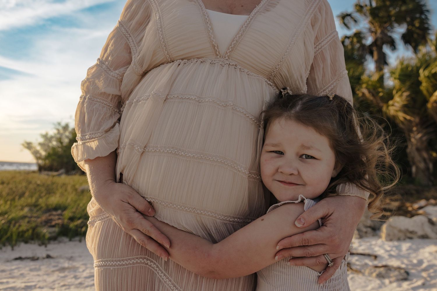 A tender moment captured at sunset on a beach as a young child hugs a pregnant belly in a flowing beige dress.
