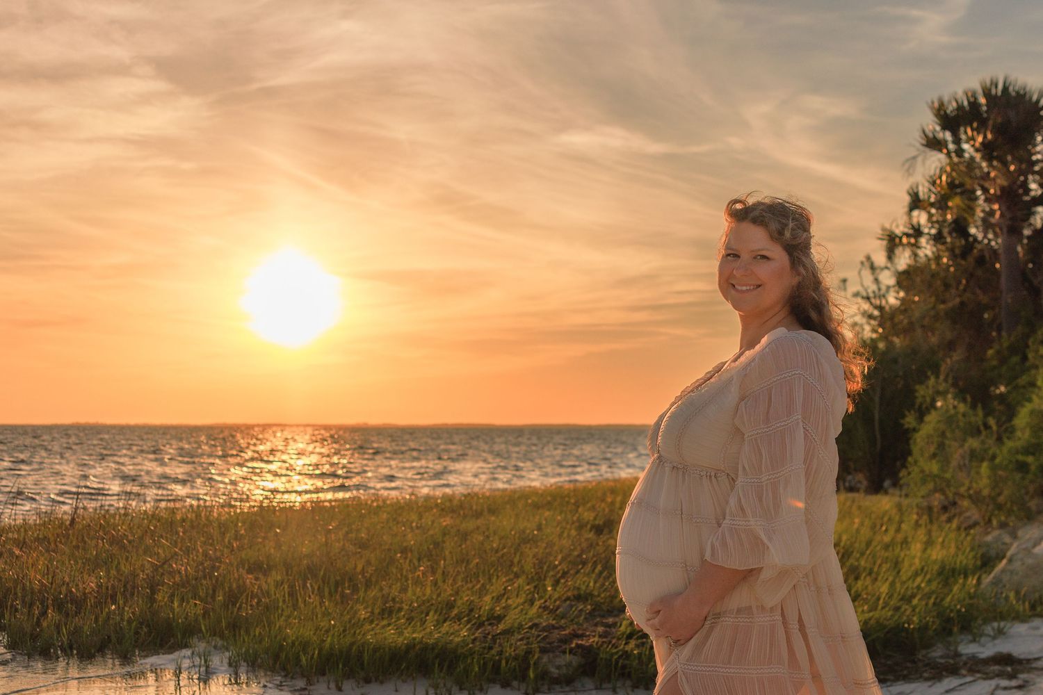 Silhouetted figure in a white dress stands on beach at sunset with glowing orange sky over ocean horizon.