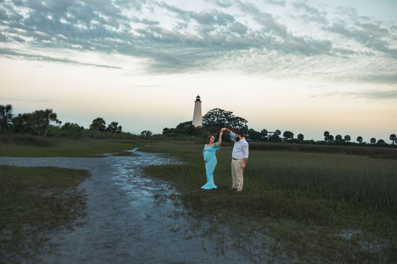 Two figures stand on a coastal path with the St Simons Lighthouse in Georgia silhouetted against a cloudy sunset sky.