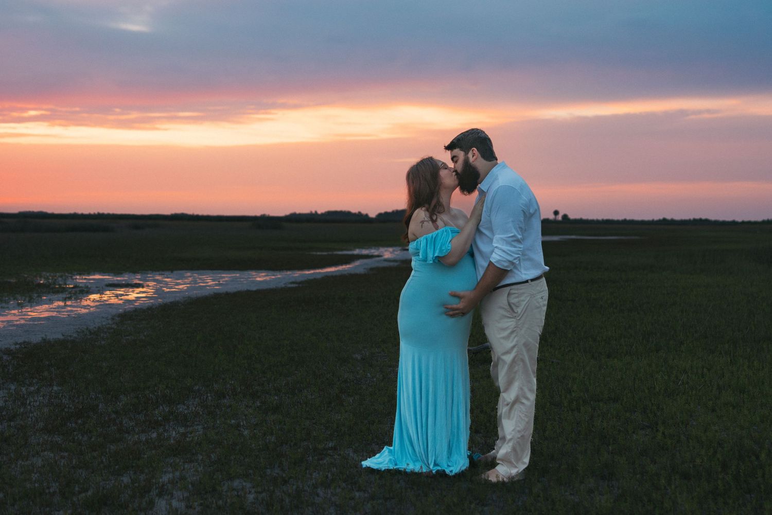 A couple in a blue maternity dress and khaki pants embrace in a grassy field during a colorful sunset.