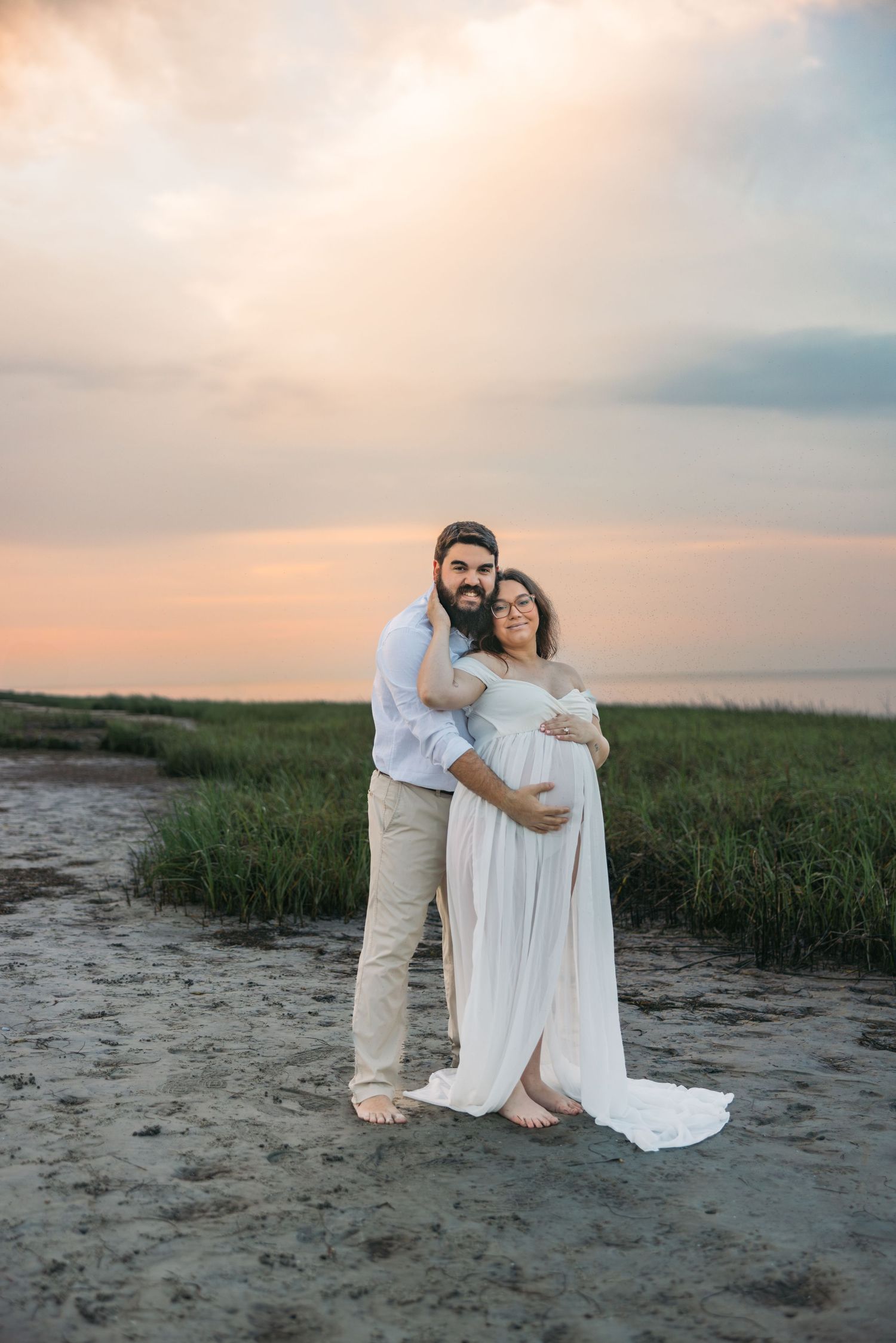 A couple standing together on a beach during sunset with soft pink clouds and green marsh grass in the background.