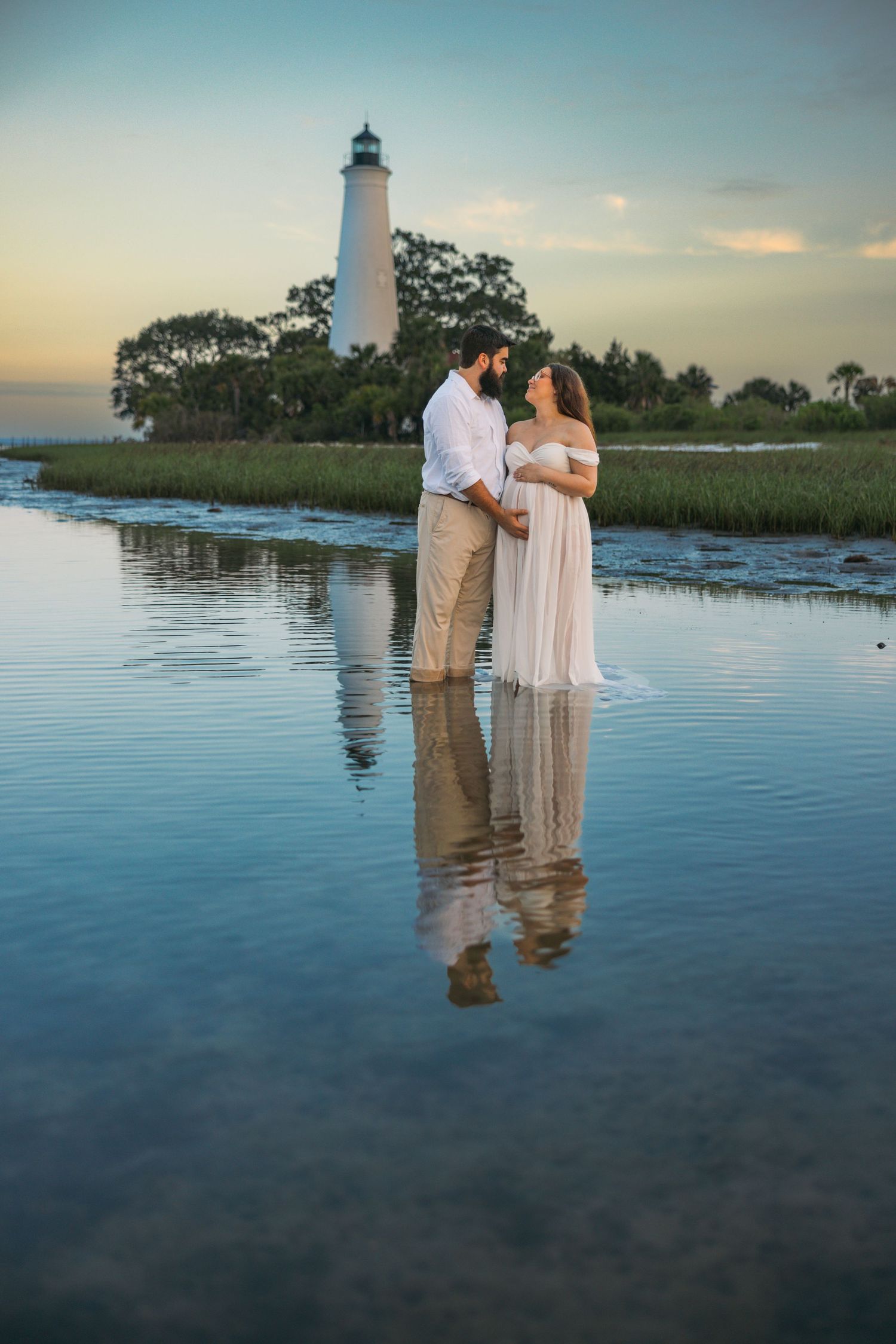 A romantic lighthouse engagement photoshoot at sunset with reflections in tranquil coastal waters.